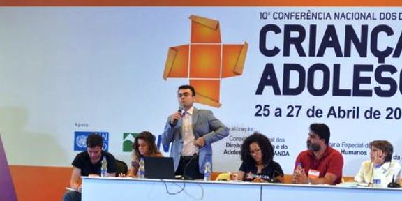 Foto de pessoas em um palco, sentados de traz de uma mesa, um homem em pé fala ao microfone.