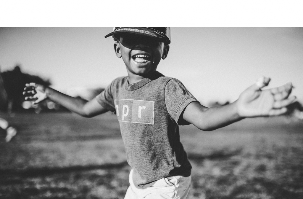 Foto em preto e branco de criança de boné sorrindo, com os braços abertos