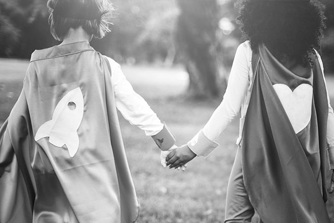 Foto em preto e branco de duas meninas de costas, com capas de super-heroínas