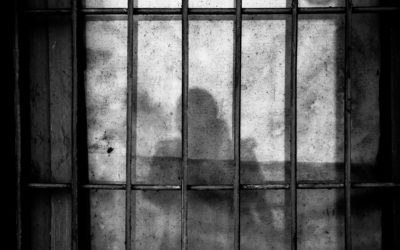 Foto em preto e branco de grades de prisão e, ao fundo, a sombra de uma pessoa