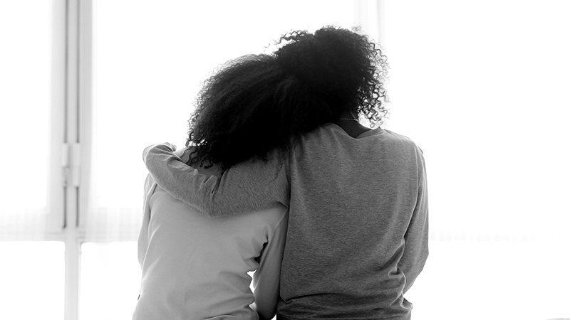 Foto em preto e branco de uma pessoa adulta abraçando uma adolescente, como se a consolasse
