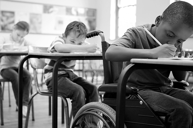 Foto em preto e branco de escola com educação inclusiva: três crianças sentadas em carteiras escrevendo, uma delas é uma pessoa com deficiência