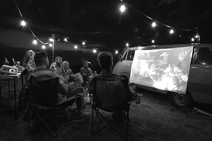 Foto em preto e branco de grupo de amigos sentados em roda assistindo um filme projetado em tela estendida na lateral de uma kombi