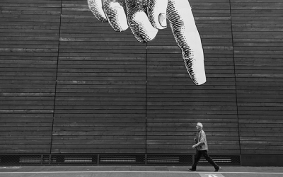 Foto em preto e branco de grafite em parede com mão apontando para pessoa que caminha na rua, em primeiro plano