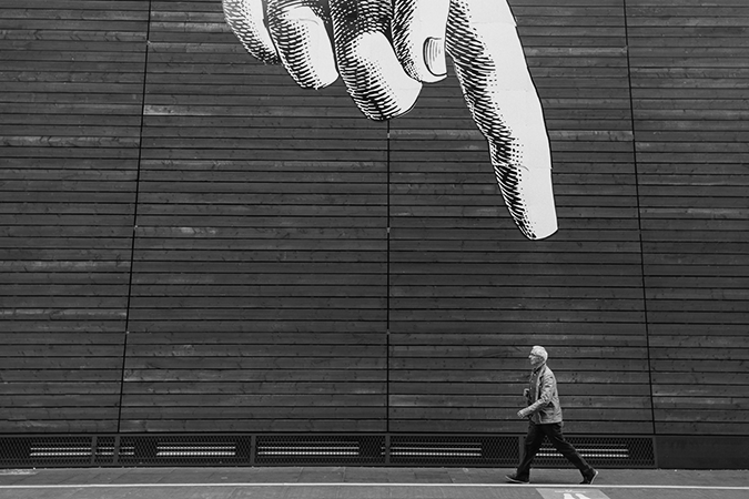 Foto em preto e branco de grafite em parede com mão apontando para pessoa que caminha na rua, em primeiro plano