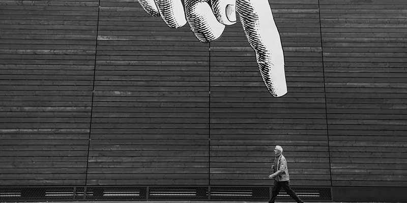 aponta Foto em preto e branco de grafite em parede com mão apontando para pessoa que caminha na rua, em primeiro plano
