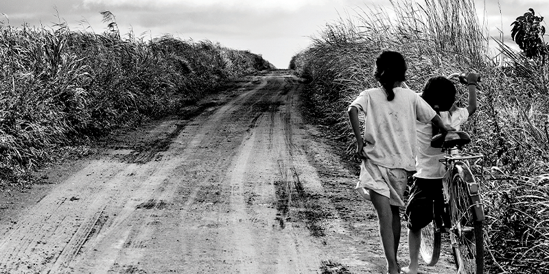 Foto em preto e branco mostra duas crianças em situação de vulnerabilidade empurrando uma bicicleta por uma estrada de terra