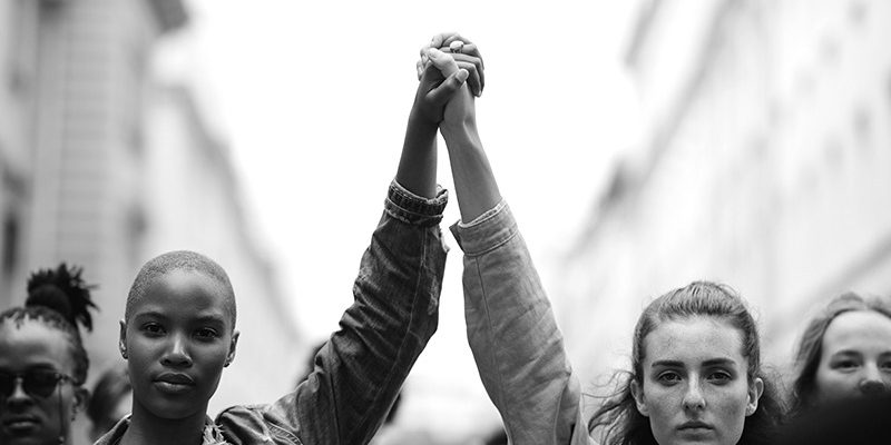 Foto em preto e branco de duas mulheres com as mãos dadas erguidas em protesto