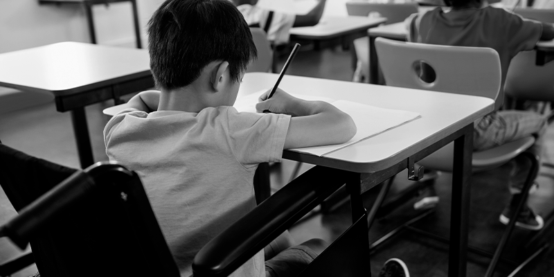 Foto em preto e branco mostra criança na escola escrevendo. Representa a necessidade da comunidade escolar estar atenta `às crianças vítimas de violência na volta às aulas