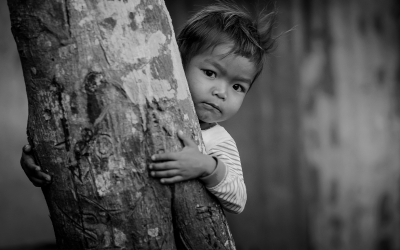 Foto em preto e branco mostra criança indígena atrás de árvore