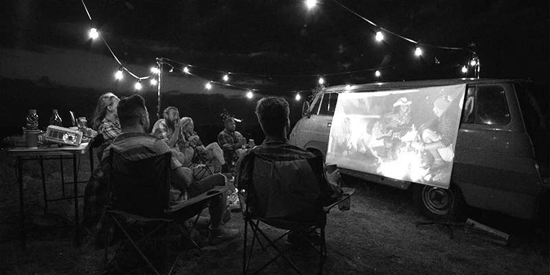 Foto em preto e branco de grupo de amigos sentados em roda assistindo um filme projetado em tela estendida na lateral de uma kombi