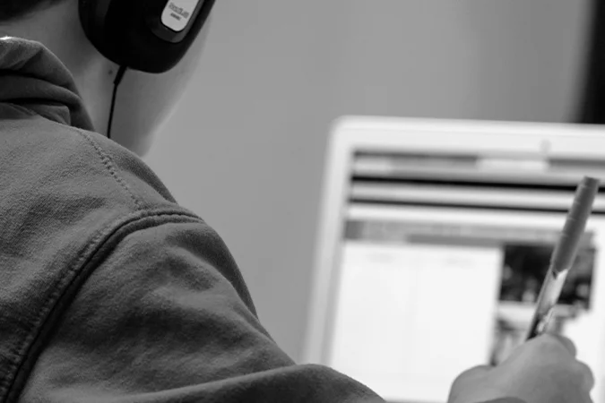 Foto em preto e branco mostra criança com fone e com uma caneta na mão acessando a internet por meio de um notebook