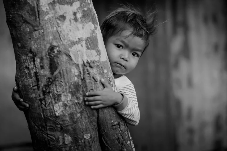 Foto em preto e branco mostra criança indígena atrás de árvore
